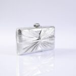 Silver rectangular clutch purse with a starburst embossed pattern and a small rounded clasp on top, photographed against a pale background with a reflective surface