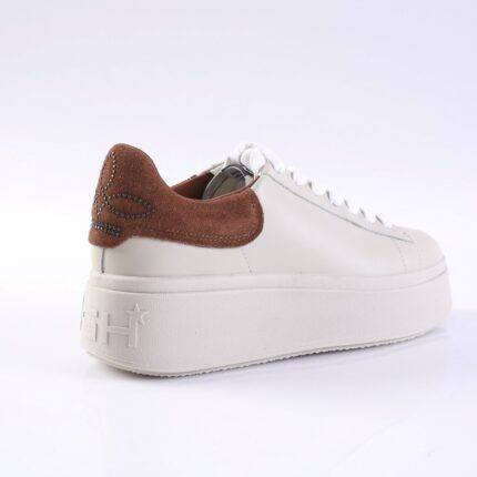 White platform sneaker with a brown suede heel counter and decorative stud pattern on the back; white laces and a chunky textured sole.