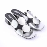Silver metallic open-toe slide sandals with oversized flower-shaped appliqués and a chunky black block heel on a white background.