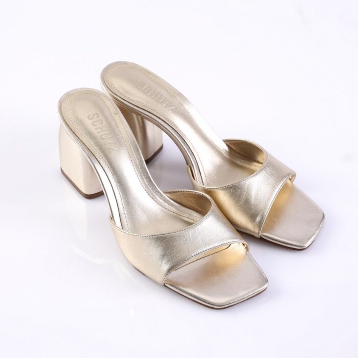 Gold metallic open-toe slide sandals with a chunky block heel on a white background, brand name SCHUTZ on the insole