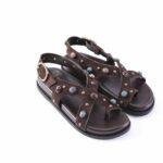 Brown suede gladiator sandals with studded straps and colorful gem accents, open-toe design on a dark sole, side buckle.