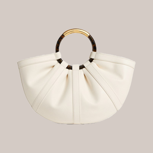 Cream crescent-shaped handbag with pleated sides and a circular tortoiseshell handle on a light beige background