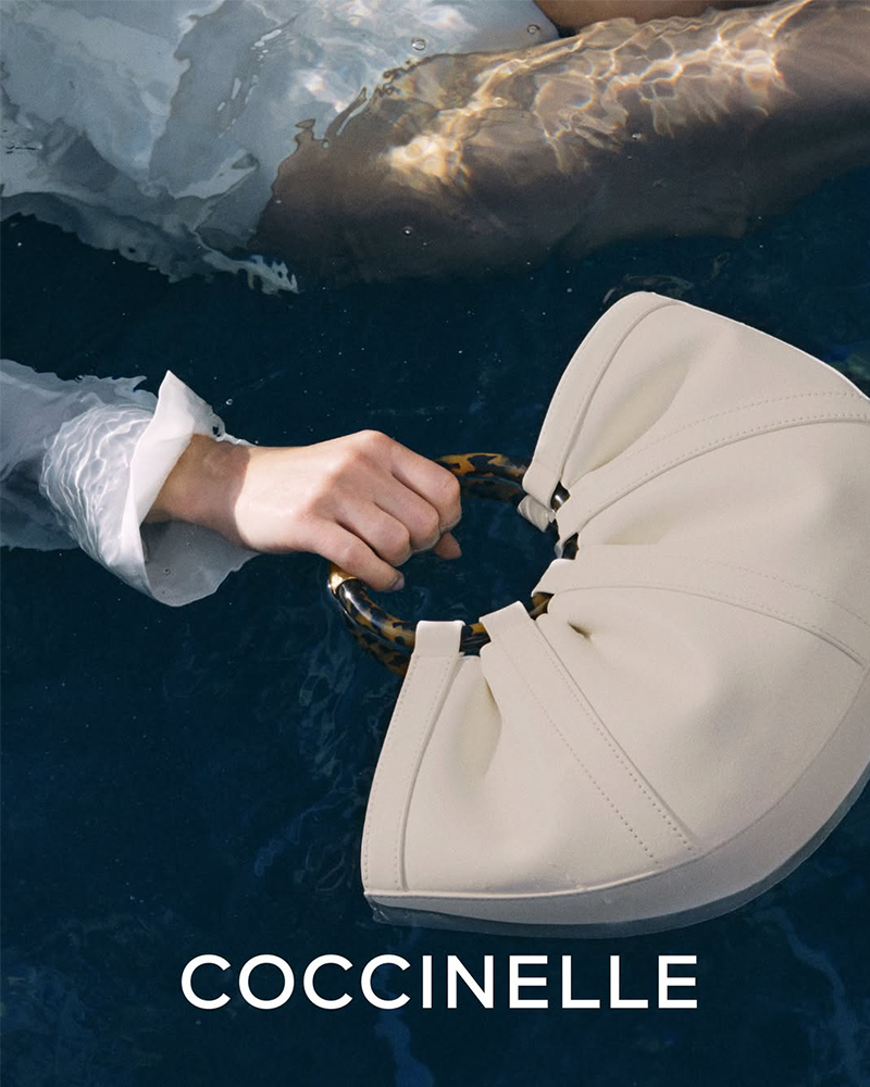 Cream handbag floating in icy water with a hand gripping its circular tortoiseshell clasp; COCCINELLE logo at bottom.