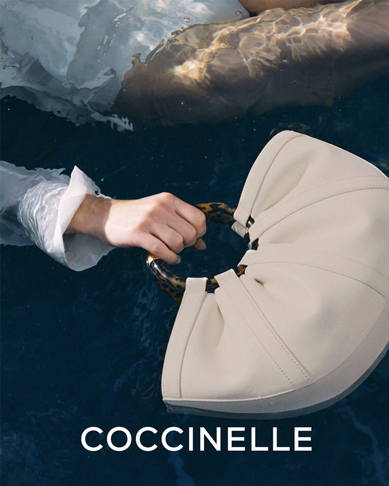 White Coccinelle leather handbag floating in dark blue water, a pale-sleeved arm reaching toward it.