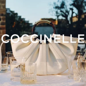 White leather handbag with a round tortoiseshell handle on a table; COCCINELLE text overlaid across the image.