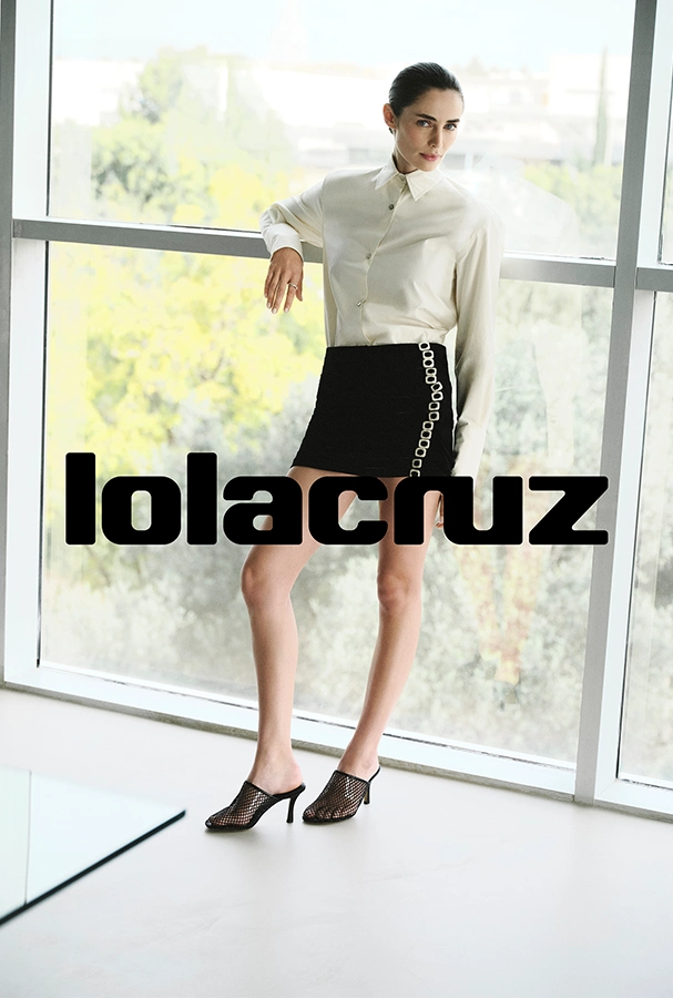 Model stands by a large window wearing a cream button-down blouse, black mini skirt with a chain detail, and mesh-heeled sandals; bold 'lolacruz' logo across the image.