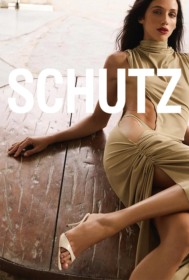 Model in a beige dress and white heels lounging against a wooden surface with a large SCHUTZ logo across the image.