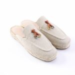 Beige canvas espadrille slip-ons with stitched seams, rope sole, and small brown leather strap with metal hardware on top.