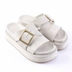 White platform sandals with two wide straps and gold square buckles on a chunky sole.